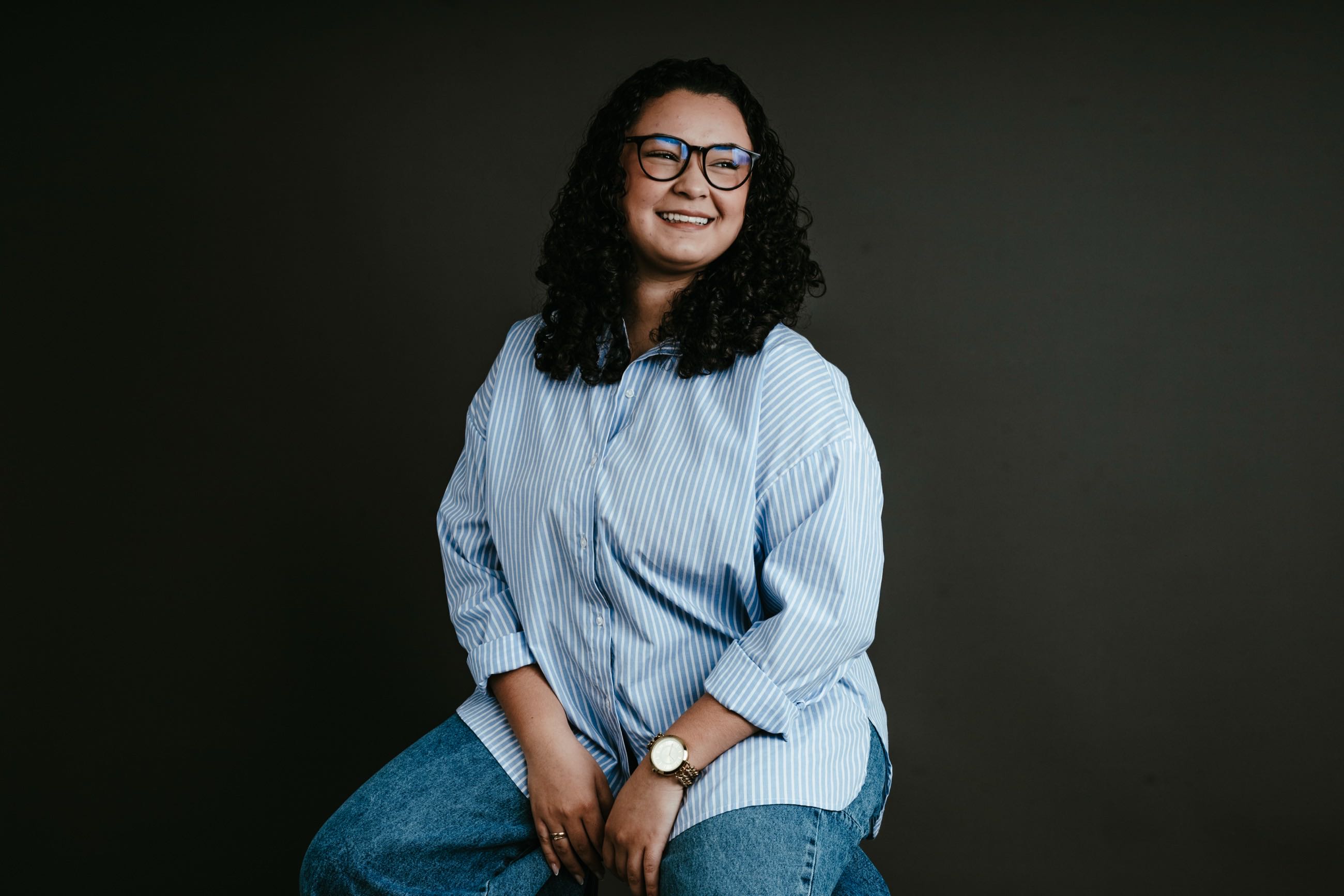Retratada sentada de camisa azul-clara, fundo escuro