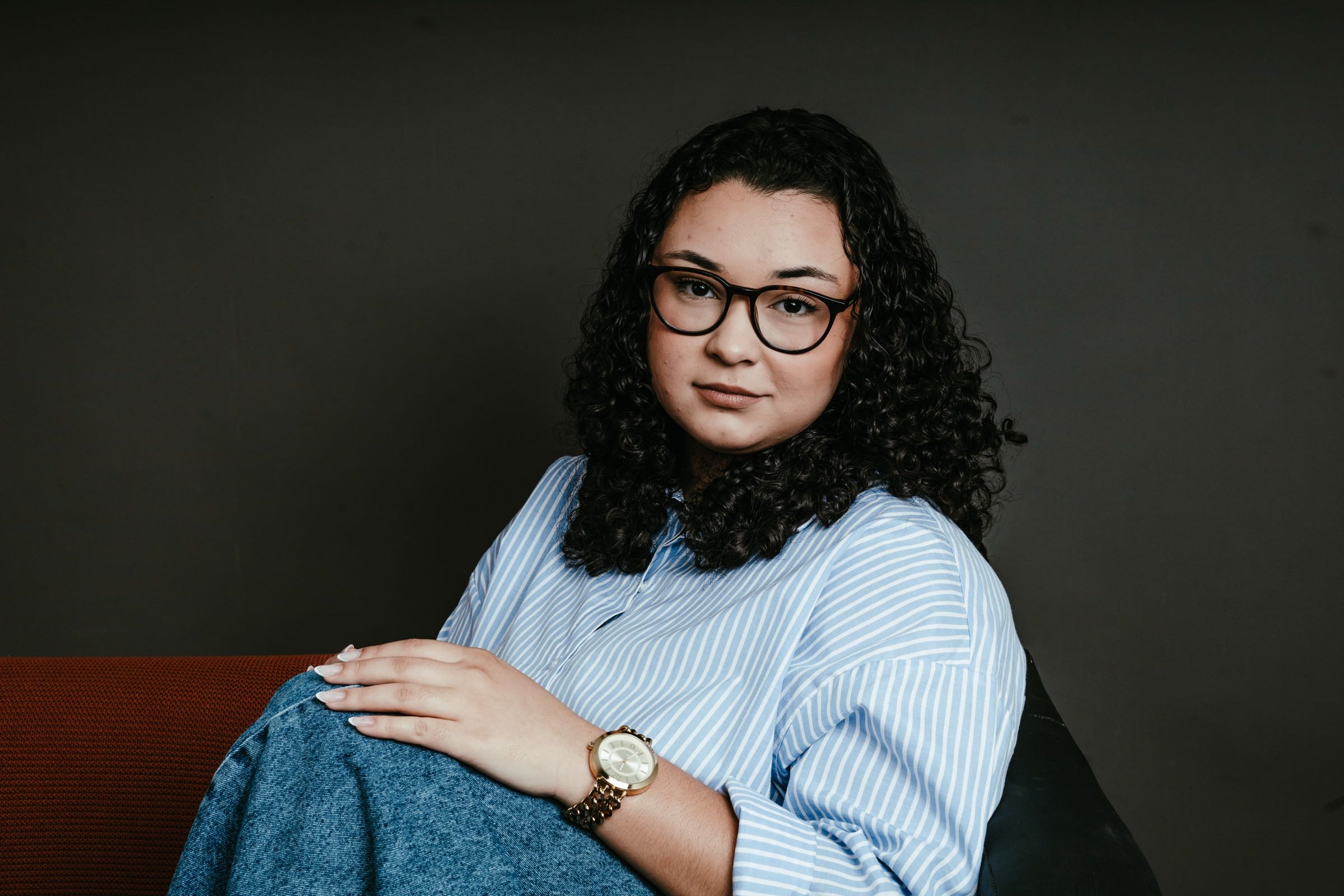 Retrato de marca pessoal: mulher de camisa listrada e óculos num sofá, fundo escuro